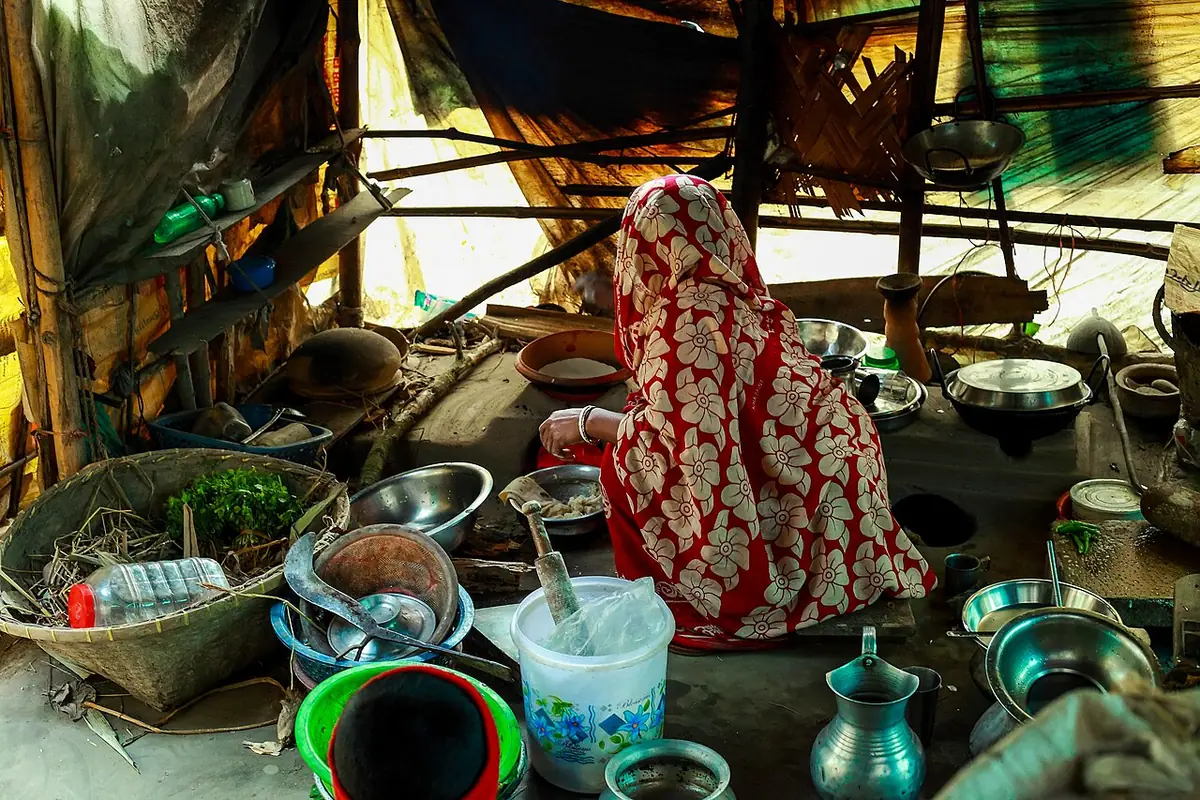 A Bangladeshi woman cooking at a clay stove in a traditional home kitchen, with steel bowls and a basket of fresh greens